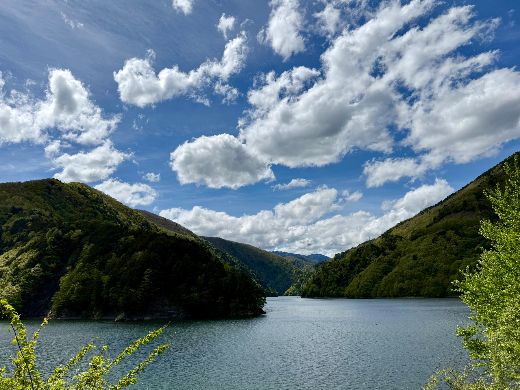 A clear, turquoise river flows through a picturesque valley with snow-capped mountains and lush greenery under a partly cloudy sky.